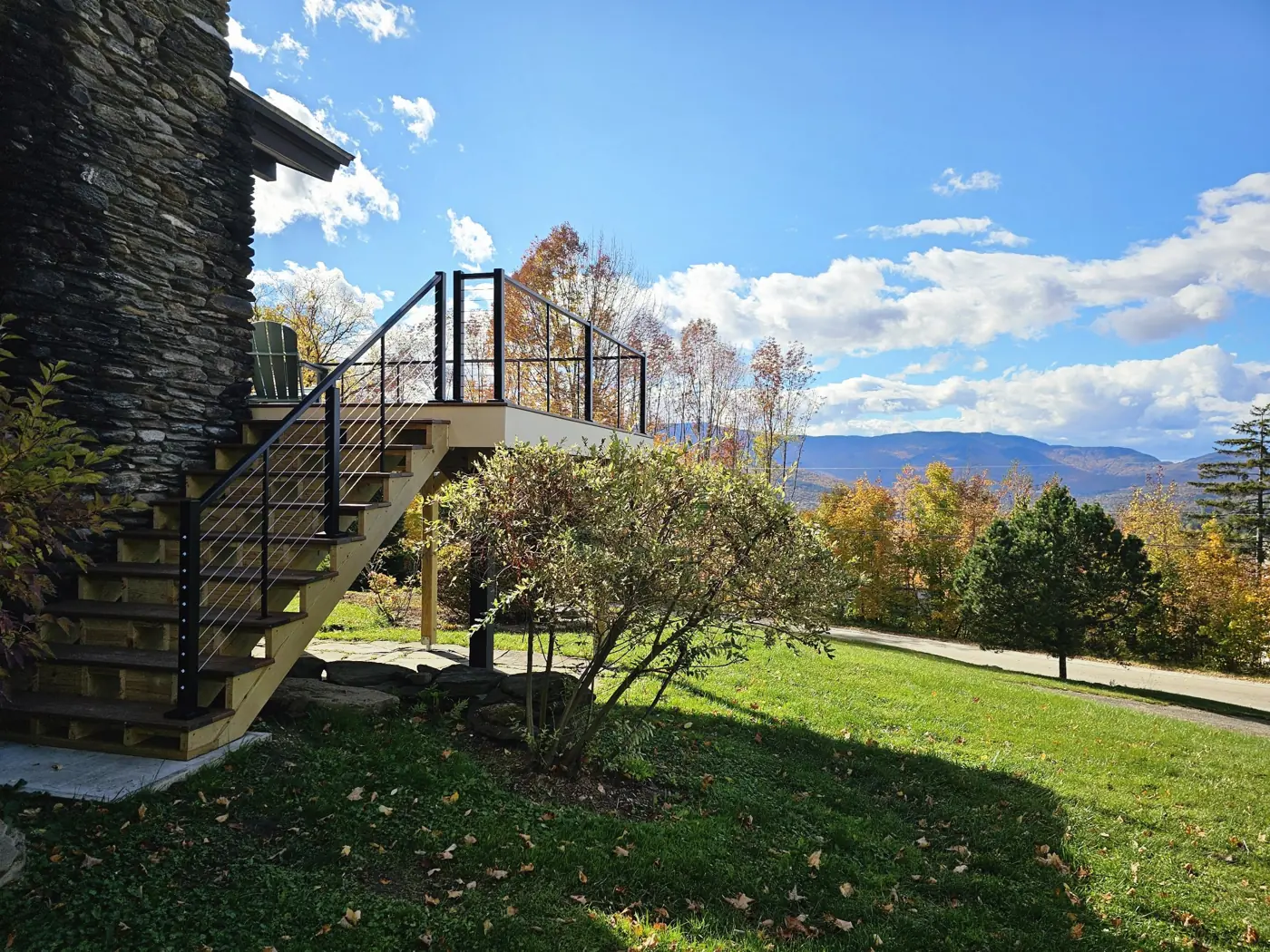 New deck stairs with cable railing beside a stone chimney, fall foliage and mountains behind